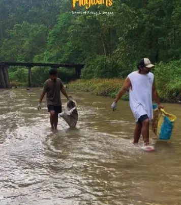 Two ploggers wading through a shallow brown river holding bags of collected plastic.